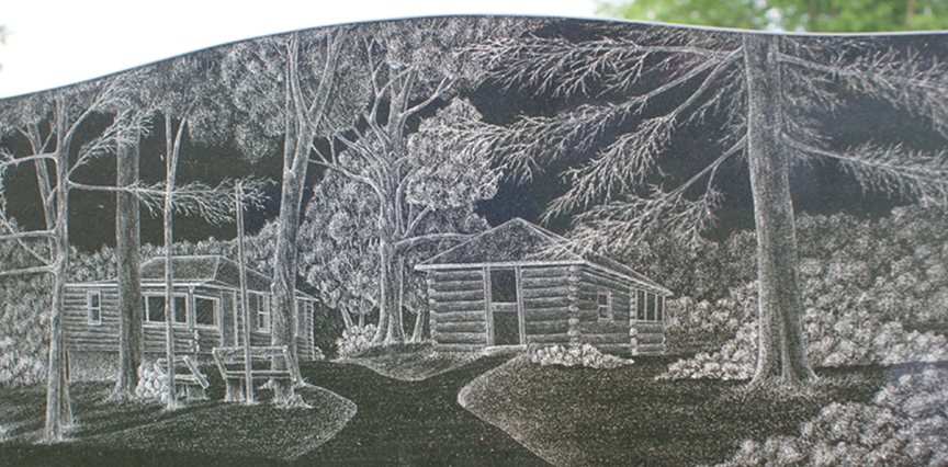 Hand etching on a shaped black granite stone of two log cabins standing among bare trees